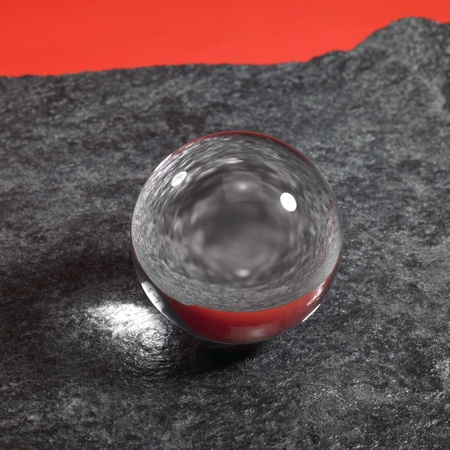a clear crystal ball on rough dark grey stone surface in front of red backの写真素材