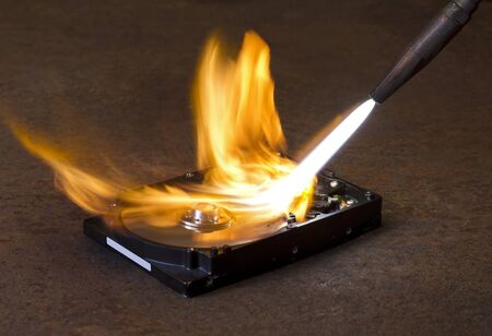 symbolic technology theme showing a welding torch tip and dashing flame burning a fixed hard disk wich is located on rusty metallic groundの写真素材
