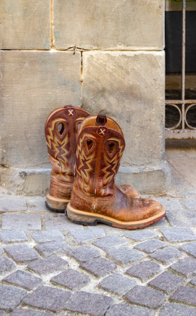 Outdoor scenery showing a pair of rundown cowboy boots on cobblestone ground in front of a sandstone wallの写真素材