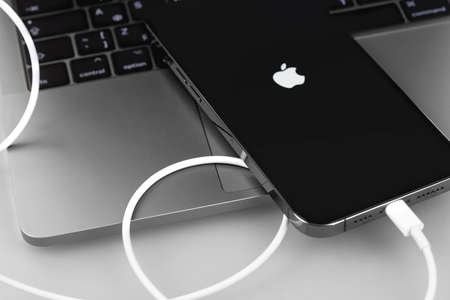 Apple iPhone smartphone connected to MacBook with Lightning cable. Apple is a multinational...