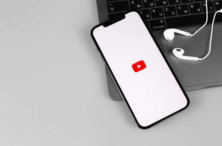 YouTube mobile app logo on screen iPhone with Macbook, Apple Earpods headphones. YouTube is a free video sharing application that anyone can watch. Moscow, Russia - December 4, 2020のeditorial素材