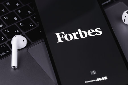 Forbes logo mobile app on screen smartphone iPhone with notebook keyboard background. Forbes is an...