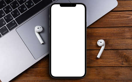 mockup smartphone iPhone 12 Pro Max with blank white screen, Macbook, AirPods top view on wooden background. Apple is a multinational technology company. Moscow, Russia - June 20, 2021のeditorial素材