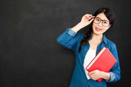 happy girl student with glasses and textbook standing in front of blackboardの写真素材