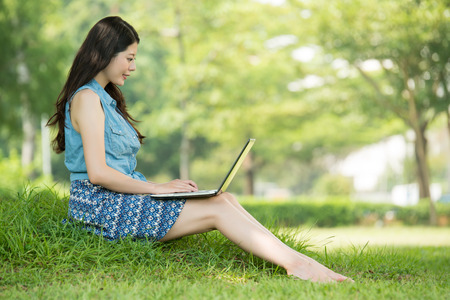 beautiful asian woman using computer sitting on grass in outdoor park. nature outdoors background. people and technology conceptの写真素材