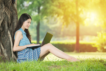 beautiful asian woman using computer sitting on grass in outdoor park. nature outdoors background. people and technology conceptの写真素材