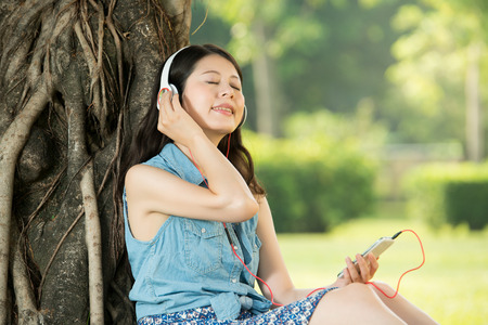 beautiful asian woman listening music sitting on grass in outdoor park. nature outdoors background. people and technology conceptの写真素材