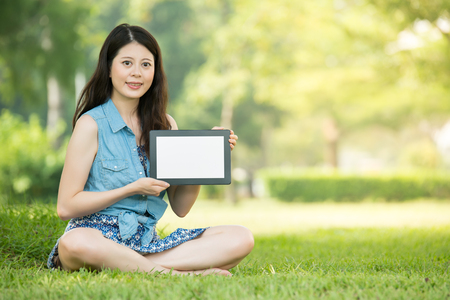 beauty asian woman presenting digital tablet sitting on grass in outdoor park. nature outdoors background. people and technology conceptの写真素材