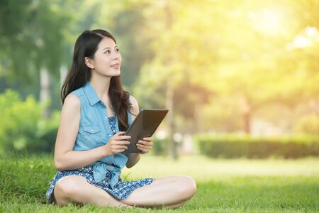 beauty asian woman using digital tablet sitting on grass in outdoor park. nature outdoors background. people and technology conceptの写真素材