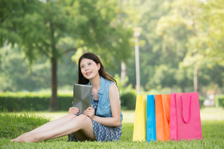 beautiful asian woman using digital tablet thinking shopping online sitting on grass. outdoors background. technology and people conceptの写真素材