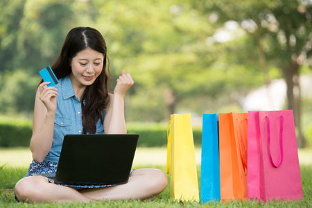 beautiful asian woman using computer shopping online with credit card sitting on grass. outdoors background. technology and people conceptの写真素材