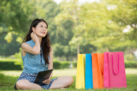 beautiful asian woman using digital tablet thinking shopping online sitting on grass. outdoors background. technology and people conceptの写真素材