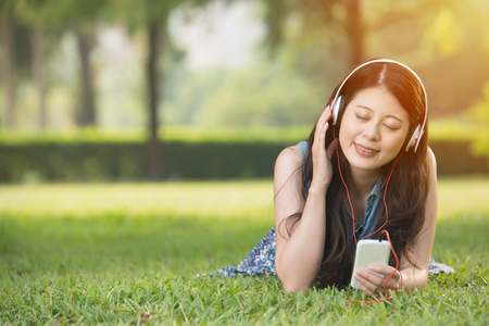 beautiful asian woman listening music with smart phone on grass in outdoor park. nature outdoors background. people and technology conceptの写真素材