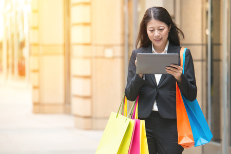 Beautiful business woman looking at her computer digital tablet while carrying shopping gift bags. young multiracial Asian womanの写真素材