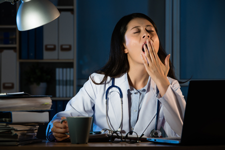 Overworked female doctor sitting at the desk and yawning at hospital. beautiful mixed race asian chinese woman model. medical and health conceptの写真素材