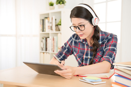 Happy student learning on line and taking notes in a digital tablet doing homework looking at pad screen in a desk at home. mixed race asian chinese model.