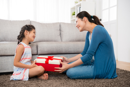 smiling daughter gave her mother a red gift box in mother's day festival holiday and sitting together in front of the living room sofa on the floor happiness.の写真素材
