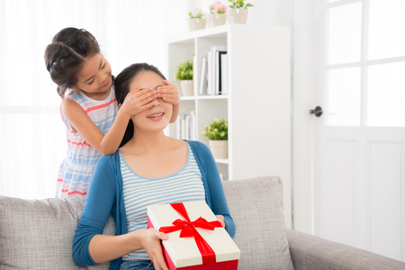 cute little girl children cover the eyes of young woman and presented a surprise at mother's day with red ribbon gift box in the living room on the sofa couch with copyspaceの写真素材