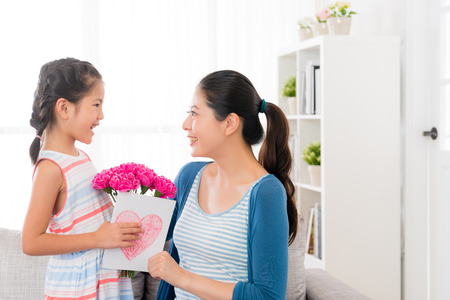 smiling asian little girl prepare a pink carnation bouquet and love card giving gift for beautiful mother at mother's day at home in the living room sofa looking each other with copyspace.の写真素材