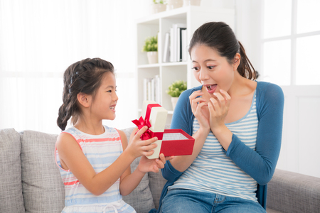 surprise mother looking at the beautiful daughter gift and open the box excited with the little girl sitting on the sofa at home in the mother's day holiday together.の写真素材