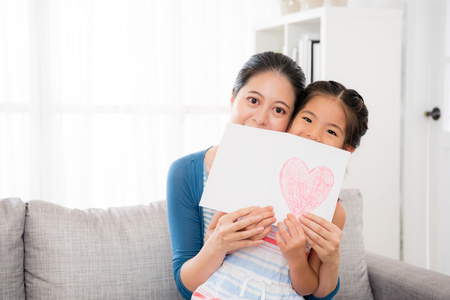 beautiful young woman with little girl embrace together sitting on couch in living room and take mother's day love card cover mouth.の写真素材