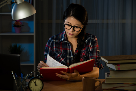 Young student at home desk reading studying at night with pile of books and coffee cup preparing exam in university education concept in dim light. mixed race asian chinese model.の写真素材