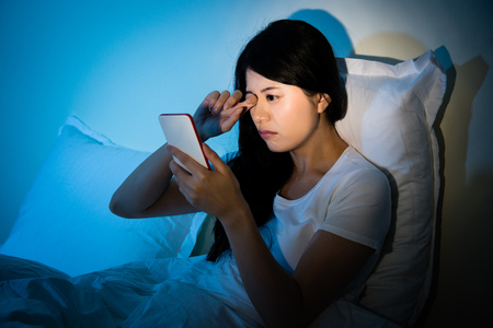 Woman rubbing her eyes feel painful with using smartphone sitting on bed sleeping at night. Mobile addict concept. mixed race asian chinese model