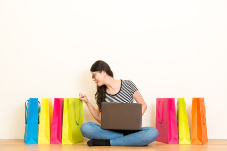 asian housewife sitting on the wood floor of the living room checking the online shopping bag goods and using the laptop with copyspace on white wall background over blank area.の写真素材