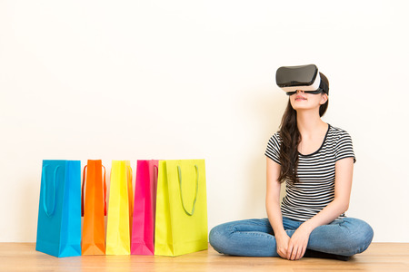 casual woman experience VR headset equipment and looking at online shopping advertising video with 3d sitting on wooden floor in the white wall background.の写真素材