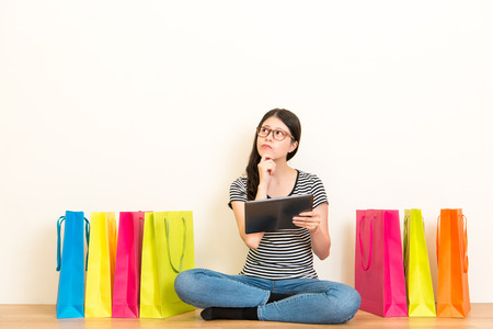 asian chinese girl worry about buying gifts for family on the festival using her mobile computer pad shopping at home online sitting on wooden floor looking at copyspace with white background.の写真素材