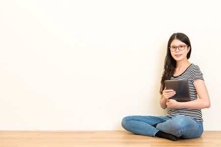 professional home teacher hold the mobile pad with blank white wall, smiling sitting on the wooden floor over copyspace. concept of studio worker.の写真素材