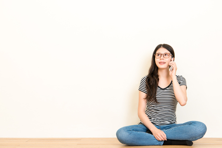 charming woman calling with cell telephone while sitting alone on wooden floor in house during free time over copyspace white wall.の写真素材
