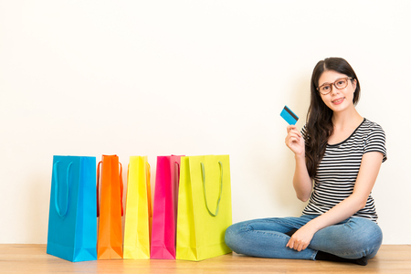 female model sitting on wooden floor ground holding credit card and showing colorful shopping bags with white background.の写真素材