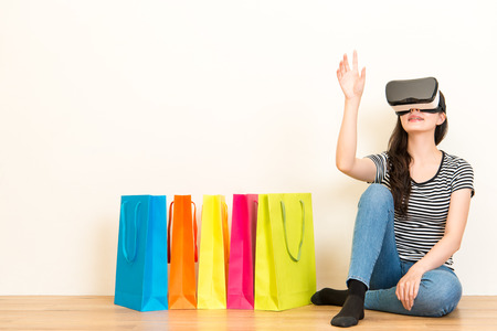 woman sitting on wooden floor with colorful shopping bags on white wall background when wearing VR device headset equipment and making grab gesture looking for goods on website.の写真素材