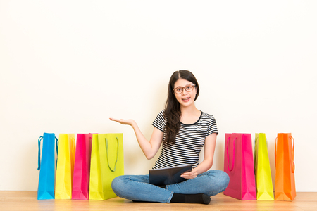 smiling woman gestures showing hands on the copyspace area sitting on the wooden floor with white wall background and holding a digital tablet with many colored shopping bags in the room.の写真素材
