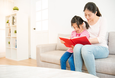 beautiful mixed race asian mother accompany with her daughter reading story book together in the living room at home. family activity concept.の写真素材