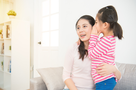 Happy cute asian kid girl whispering the secret to her laughing happy mother in ear with fun face in the living room at home. family activity concept.