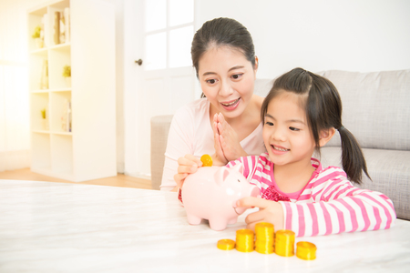 cute happy asian girl kid putting money coins into piggy bank with her mother together in the living room at home. family activity concept.の写真素材
