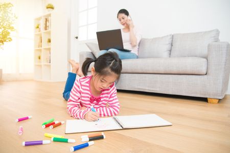 kids girl lying down on wooden floor drawing on sketchbook and mother working with computer talking on the phone on background in the living room at home. family activity conceptの写真素材