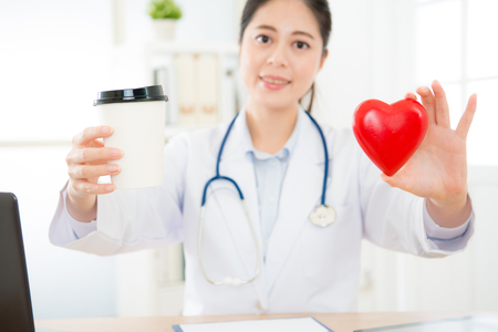 coffee cup and heart model showing caffeine causing people getting palpitations problem concept with selective focus photo of blur smiling female doctor in background.の写真素材