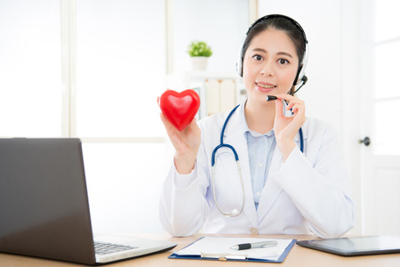 smiling female doctor using headphone to conversation at clinic office with cardiology patient using online technology service system.の写真素材
