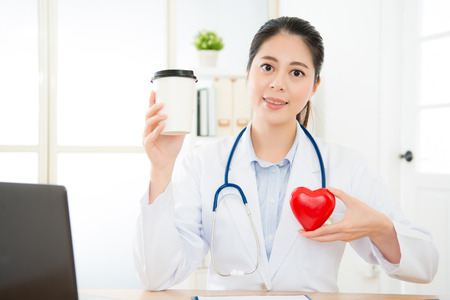 smiling young female cardiology doctor face to camera on working desk and holding coffee cup showing caffeine causing palpitations problem concept.の写真素材