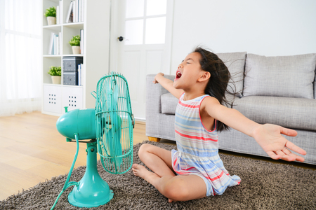 pretty happy female kid opening arms enjoying cool wind from electric fan and sitting on floor in living room at summer vacation.の写真素材