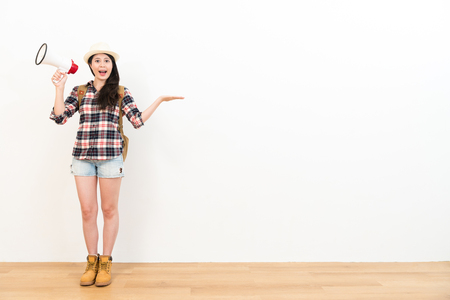 sweet pretty female traveler making displaying gesture showing white wall background and wearing travel clothing holding megaphone standing on wooden floor looking at camera.の写真素材