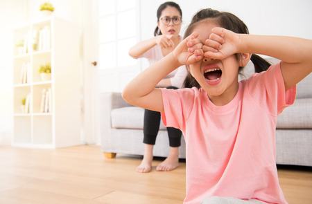 the sad little children girl sitting on the wooden floor crying loudly when the mother pointing to her sitting on sofa in the living room angry curse on the morning of the holiday.