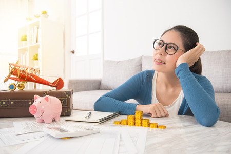 beautiful woman sitting in home living room imagine foreign travel with piggy bank gold coin and daydreaming when she recorded in family bill cost account expenses.の素材 [FY31083497276]