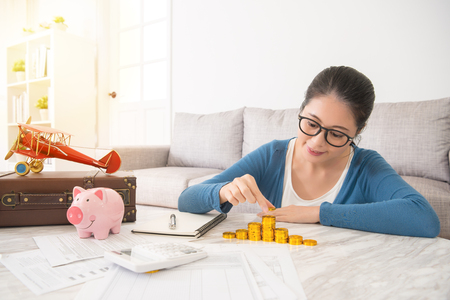 asian chinese woman opening pink piggybank deposit savings and calculate the amount of gold coins on the bill table in the living room at home for travel vacation with her friends.の写真素材