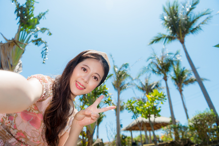 happy beautiful lady showing victory cheerful gesture and looking at camera smiling taking selfie photo with tropical coconut tree landscape.の写真素材