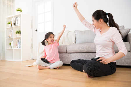 family holiday lifestyle of happy little girl daughter and elegant beautiful mom sitting on wooden floor raised fist hand gesture to celebrate winning video games  and look at each other.の写真素材