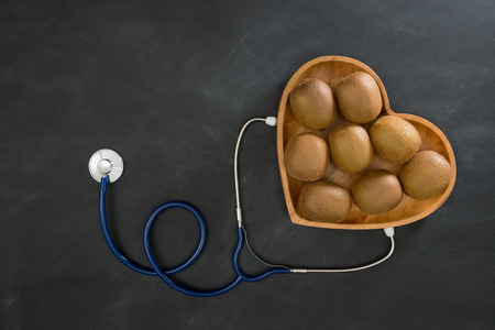 high angle photo of ripe fresh kiwi fruit group in wooden heart shaped plate with stethoscope equipment isolated on black chalkboard.の写真素材